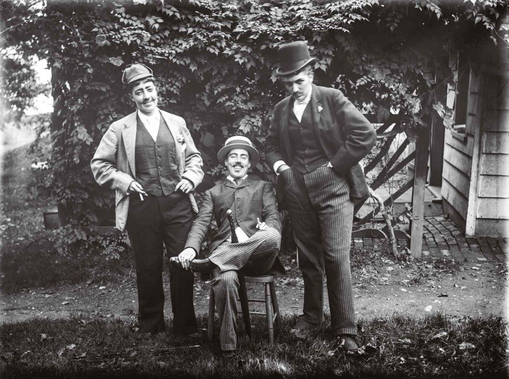 Julia Martin, Julia Bredt et moi-même en habit (1891). L’un des clichés les plus connus d’Alice Austen, qui pose au centre. Cette mise en scène des stéréotypes de la masculinité illustre son sens de l’humour subversif : elle tient, de manière suggestive, un parapluie entre les jambes.Crédit photo : Collection of Historic Richmond Town